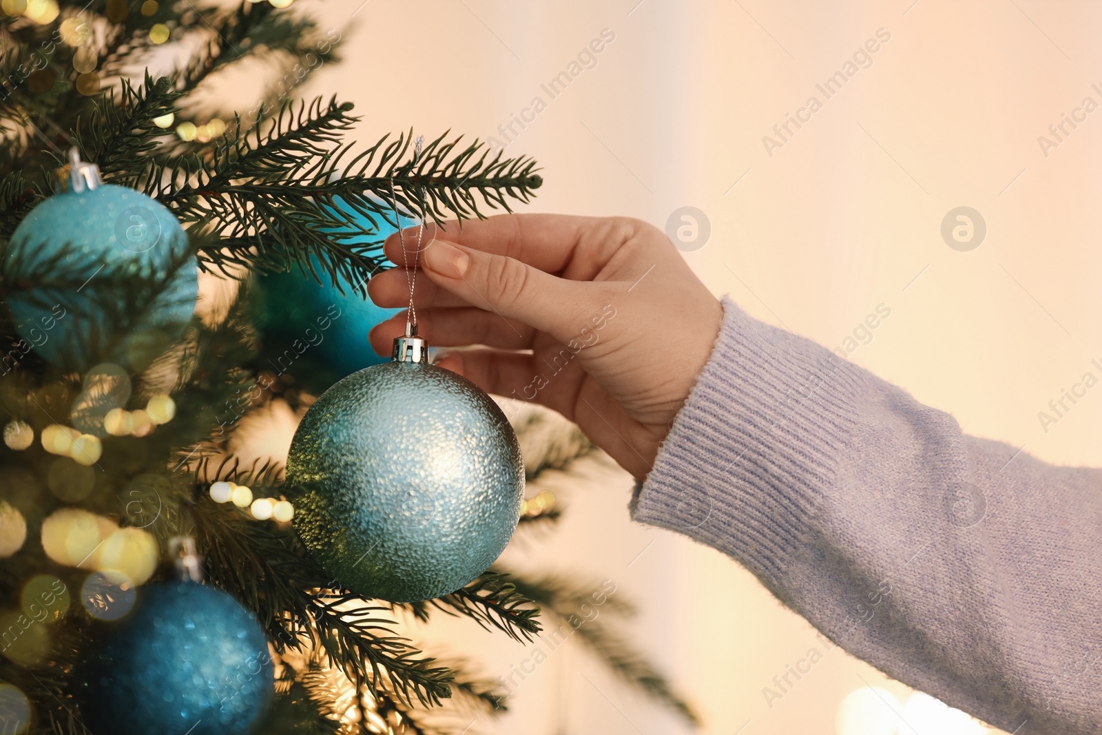 Photo of Woman decorating Christmas tree with light blue festive ball on blurred background, closeup