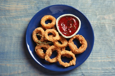 Plate with homemade crunchy fried onion rings and tomato sauce on wooden background, top view