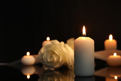 White rose and burning candles on black mirror surface in darkness, closeup with space for text. Funeral symbols