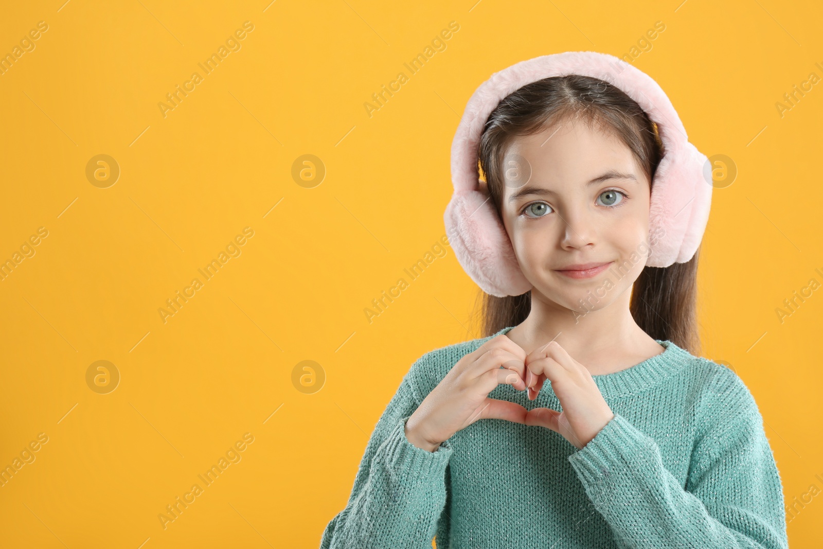 Photo of Cute girl in stylish earmuffs making heart with hands on yellow background. Space for text