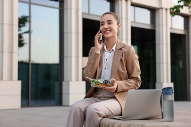 Photo of Happy businesswoman with container of salad talking on smartphone during lunch on bench outdoors