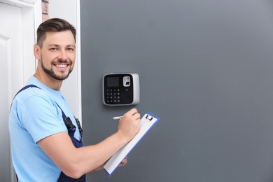 Photo of Male technician with clipboard near installed alarm system indoors