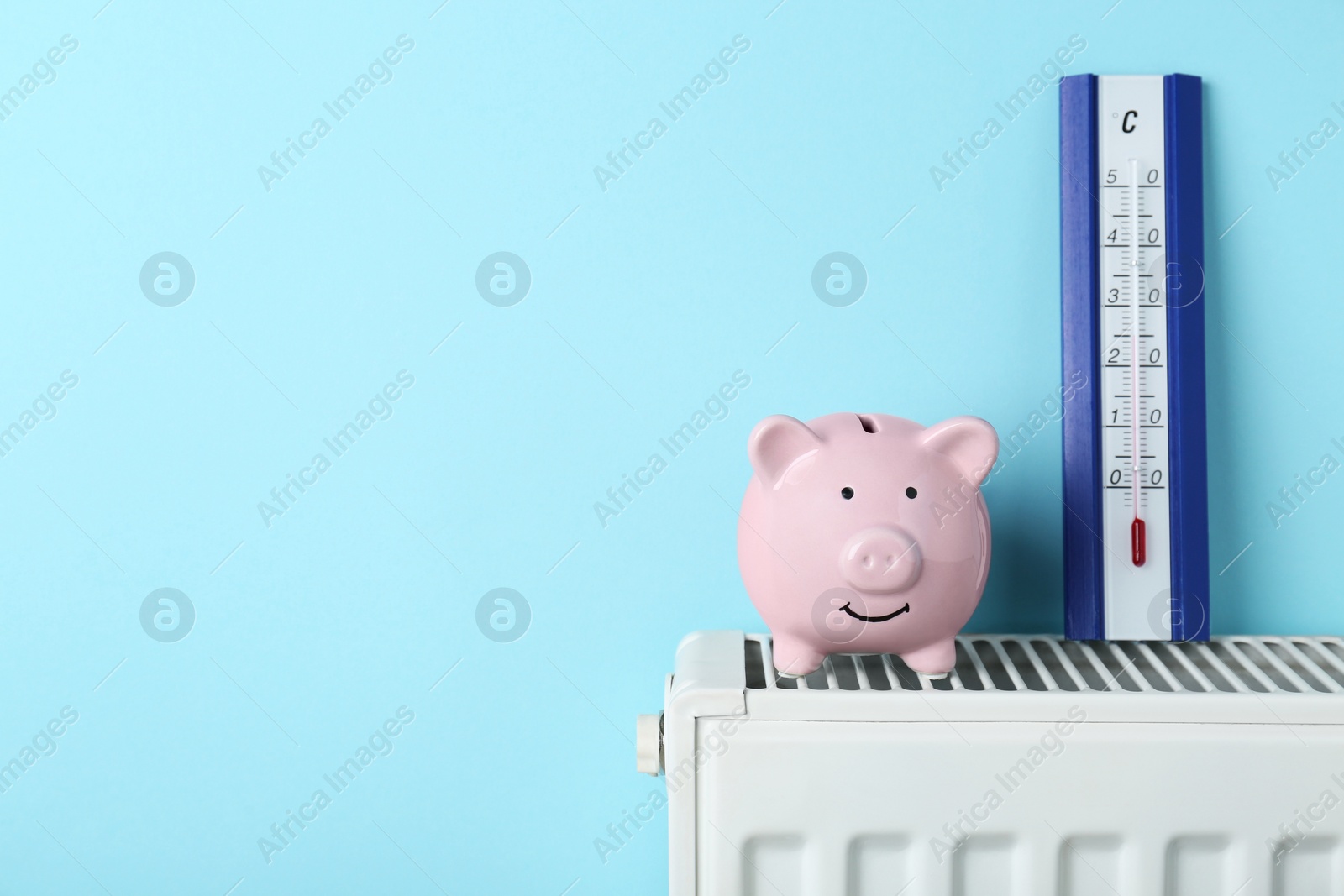 Photo of Piggy bank and thermometer on heating radiator against light blue background, space for text