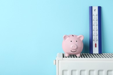 Photo of Piggy bank and thermometer on heating radiator against light blue background, space for text