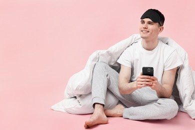 Photo of Happy man in pyjama with sleep mask, blanket and smartphone on pink background, space for text