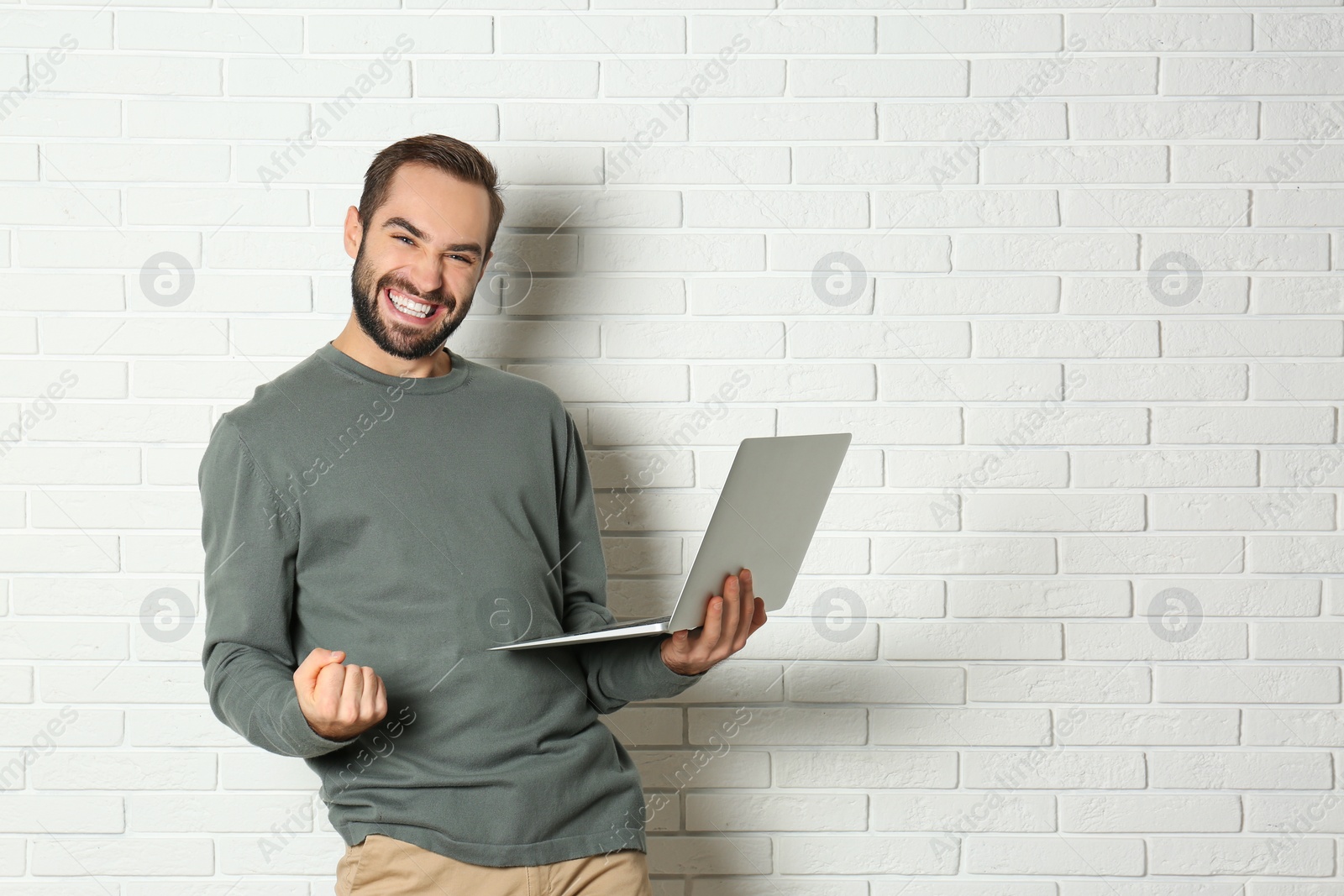 Photo of Emotional young man with laptop celebrating victory near brick wall. Space for text