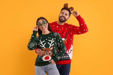 Photo of Happy young couple in Christmas sweaters, reindeer headband and funny glasses on orange background