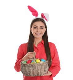 Beautiful woman in bunny ears headband holding basket with Easter eggs on white background