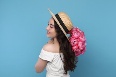Photo of Beautiful young woman in straw hat with bouquet of pink peonies against light blue background