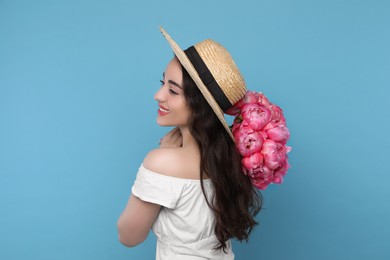 Beautiful young woman in straw hat with bouquet of pink peonies against light blue background