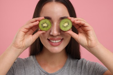 Woman covering eyes with halves of kiwi on pink background