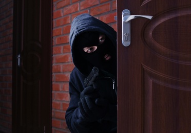 Masked man with gun spying behind open door indoors. Criminal offence