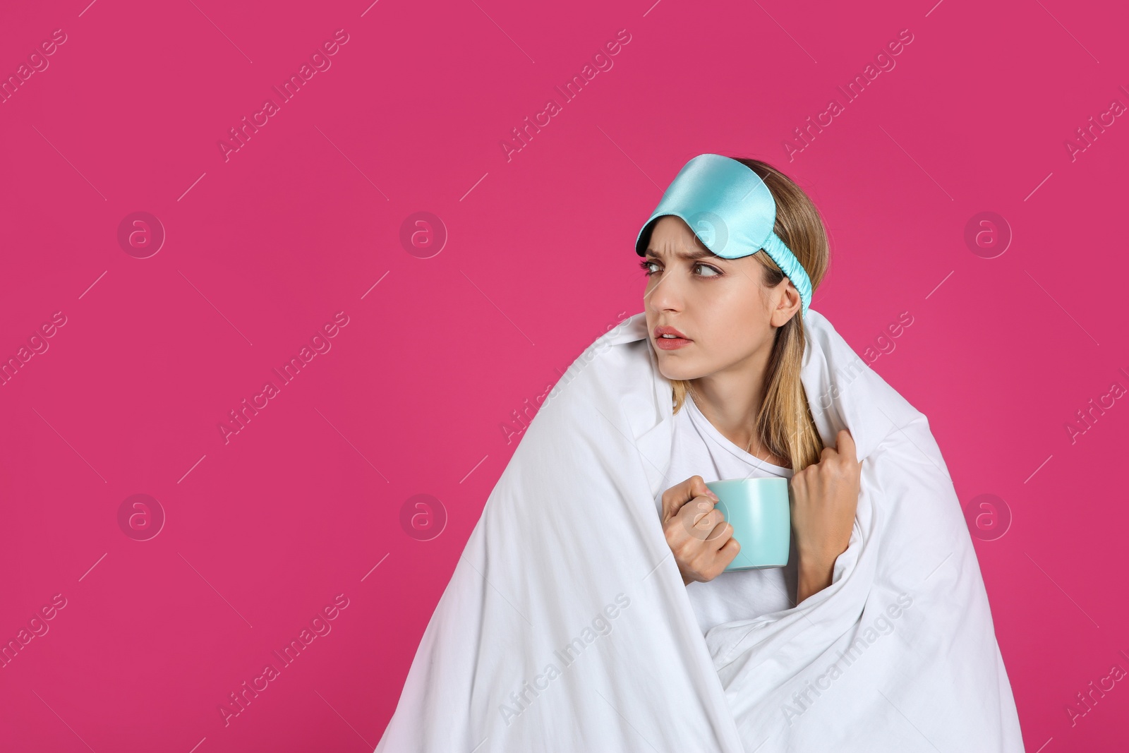 Photo of Sad young woman in sleeping mask wrapped with blanket holding cup on pink background. Space for text