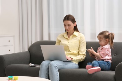 Photo of Mother working remotely on laptop while her daughter playing with smartphone at home