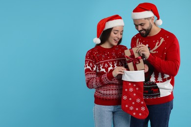 Happy young couple in Christmas sweaters and Santa hats taking gift from stocking on light blue background. Space for text
