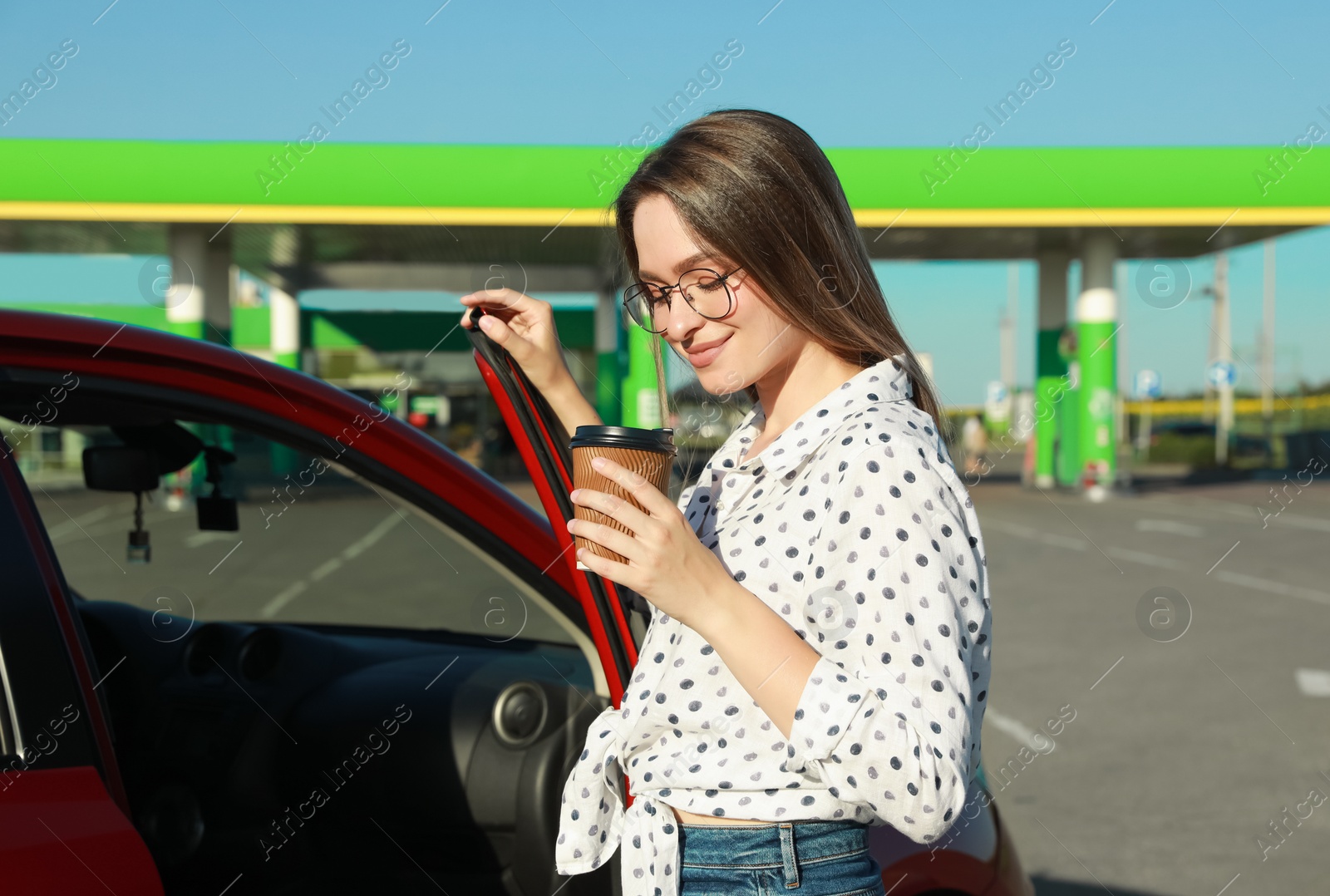 Photo of Beautiful young woman with coffee opening car door at gas station