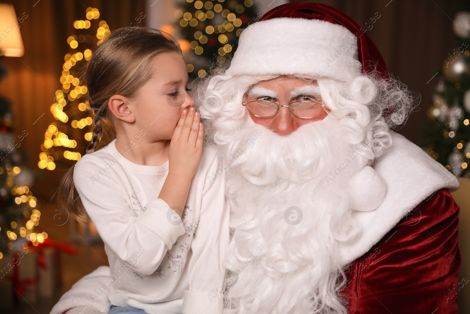 Photo of Little girl whispering in Santa Claus' ear near Christmas tree indoors