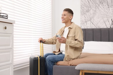 Photo of Smiling guest with suitcase and smartphone on bed in stylish hotel room