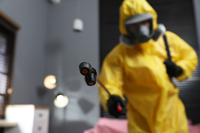 Photo of Pest control worker in protective suit indoors, focus on insecticide sprayer