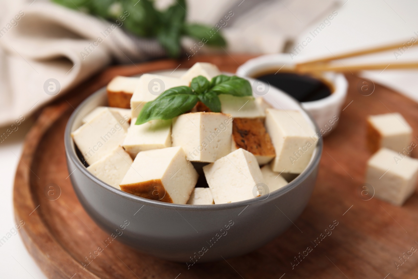 Photo of Bowl of smoked tofu cubes, soy sauce and basil on wooden tray, closeup