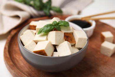 Photo of Bowl of smoked tofu cubes, soy sauce and basil on wooden tray, closeup