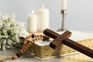 Wooden cross, Bible, rosary beads, flowers and church candles on light table, closeup