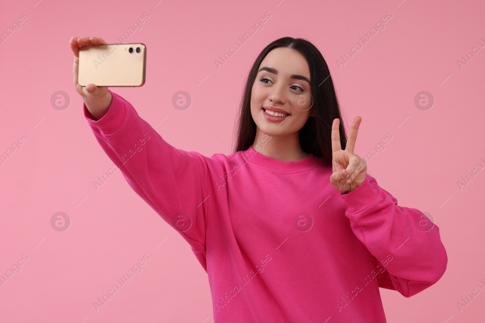 Photo of Smiling young woman taking selfie with smartphone and showing piece sign on pink background, space for text