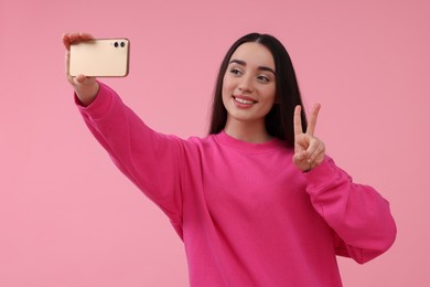 Photo of Smiling young woman taking selfie with smartphone and showing piece sign on pink background, space for text