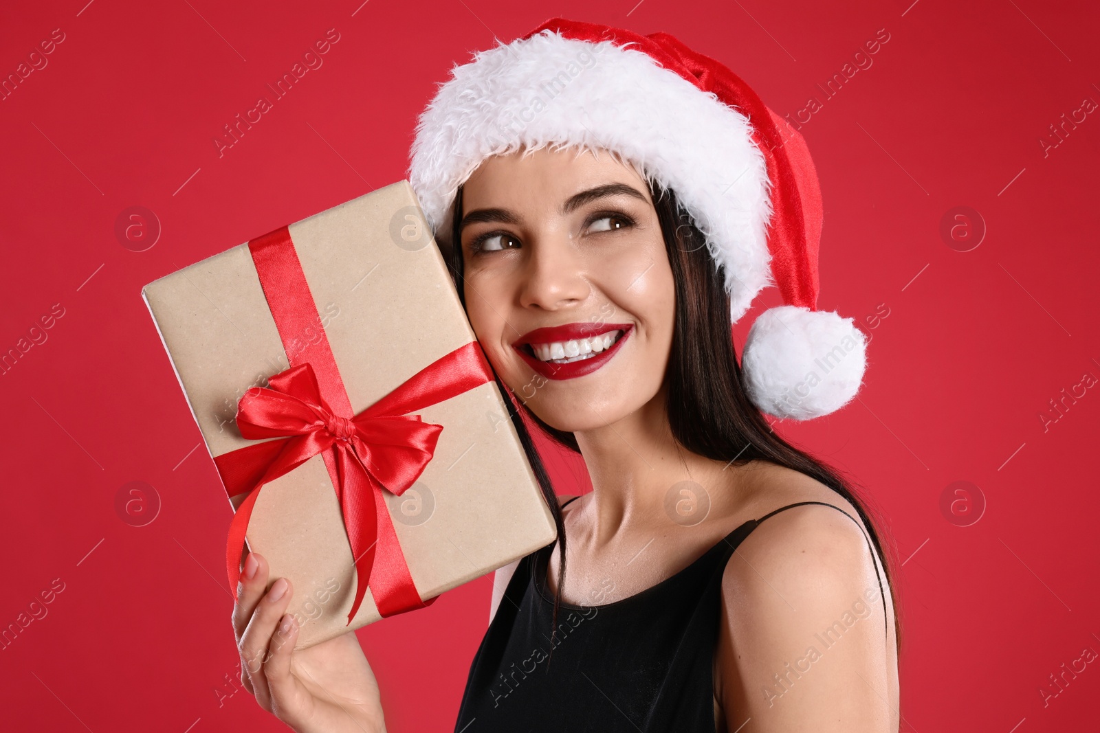 Photo of Woman in black dress and Santa hat holding Christmas gift on red background