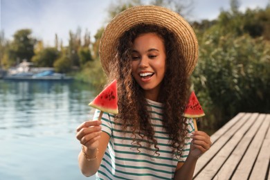 Photo of Beautiful young African American woman with pieces of watermelon on wooden pier near river