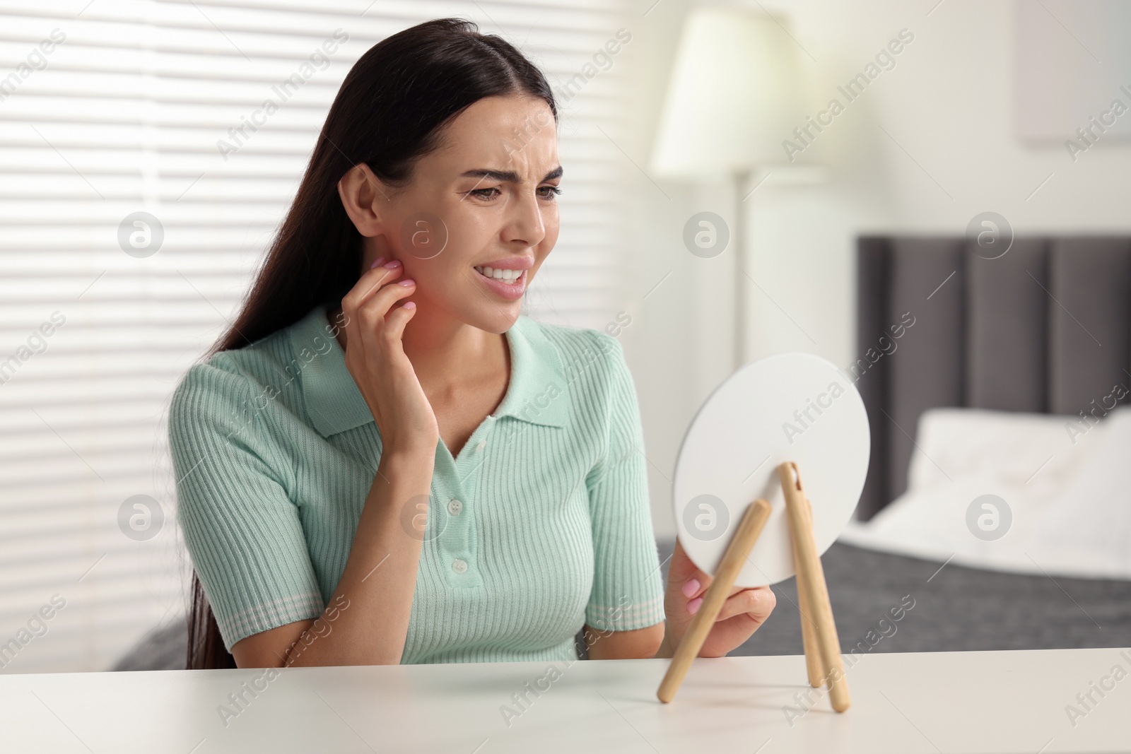 Photo of Suffering from allergy. Young woman looking in mirror and scratching her face at white table indoors