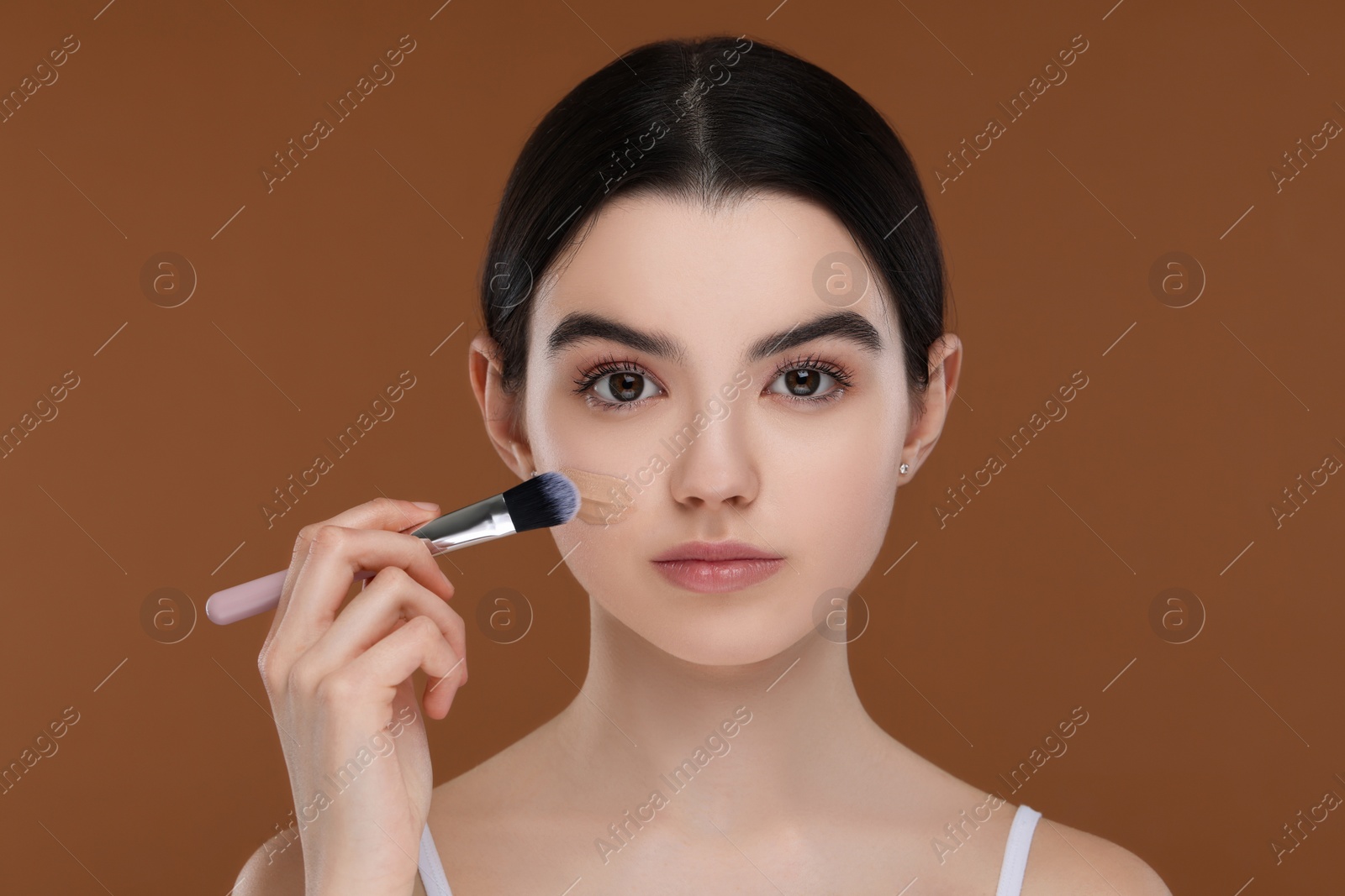 Photo of Teenage girl applying foundation on face with brush against brown background