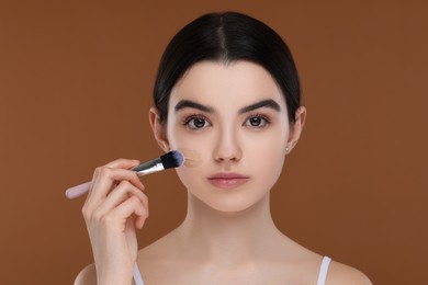 Teenage girl applying foundation on face with brush against brown background