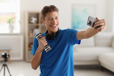 Photo of Smiling sports blogger with dumbbell streaming online fitness lesson with smartphone at home