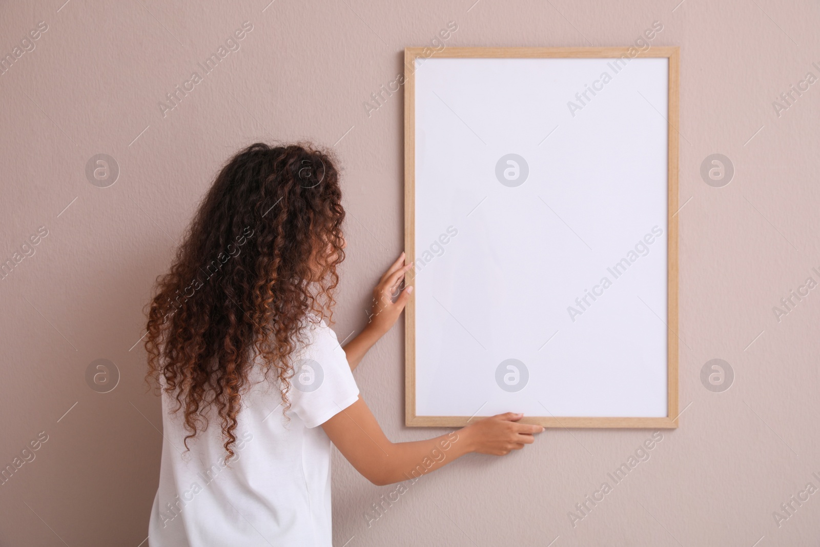 Photo of African American woman hanging empty frame on pale rose wall, back view. Mockup for design