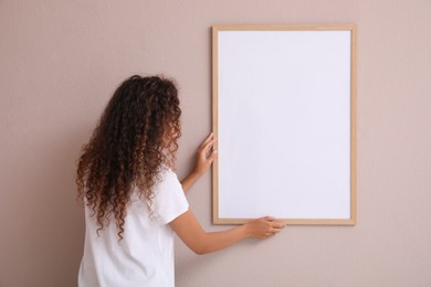 Photo of African American woman hanging empty frame on pale rose wall, back view. Mockup for design