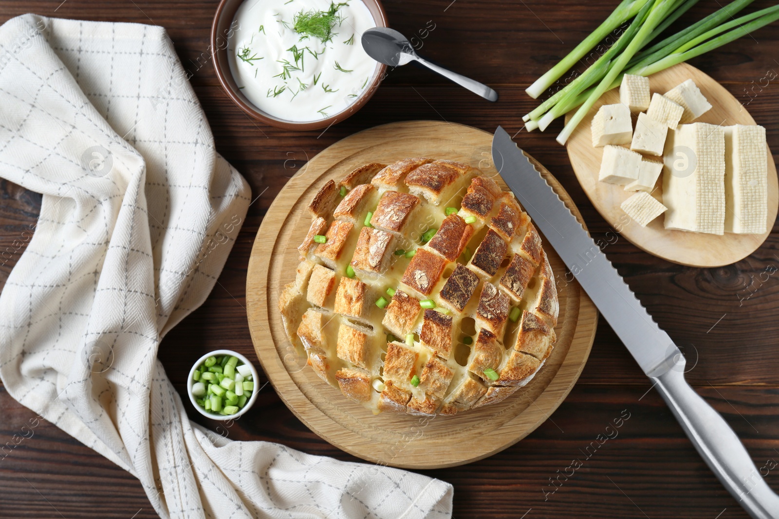 Photo of Freshly baked bread with tofu cheese, green onions, sauce and knife on wooden table, flat lay