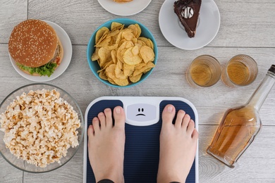 Photo of Woman standing on scales surrounded by different food and alcohol after party indoors, top view