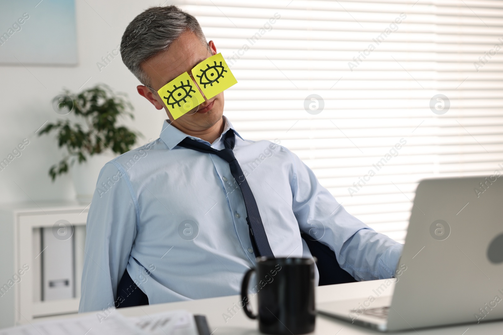 Photo of Man with fake eyes painted on sticky notes snoozing at workplace in office