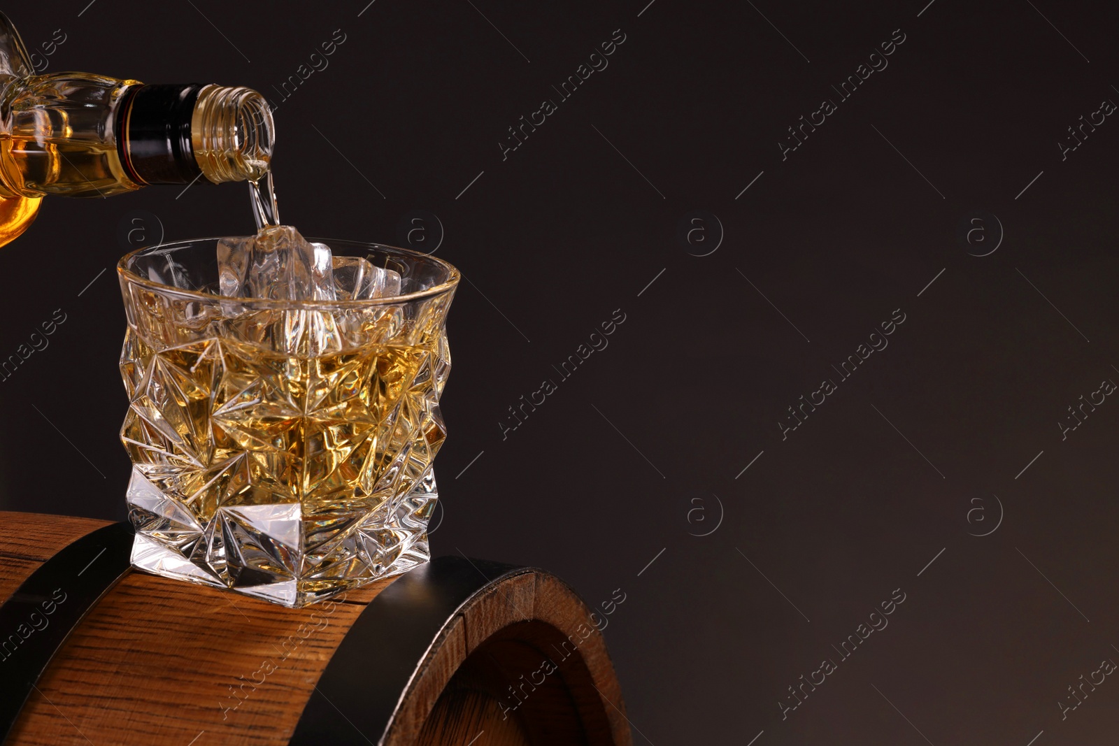 Photo of Pouring whiskey from bottle into glass on wooden barrel against dark background, closeup. Space for text