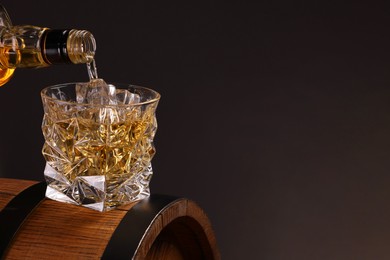 Photo of Pouring whiskey from bottle into glass on wooden barrel against dark background, closeup. Space for text