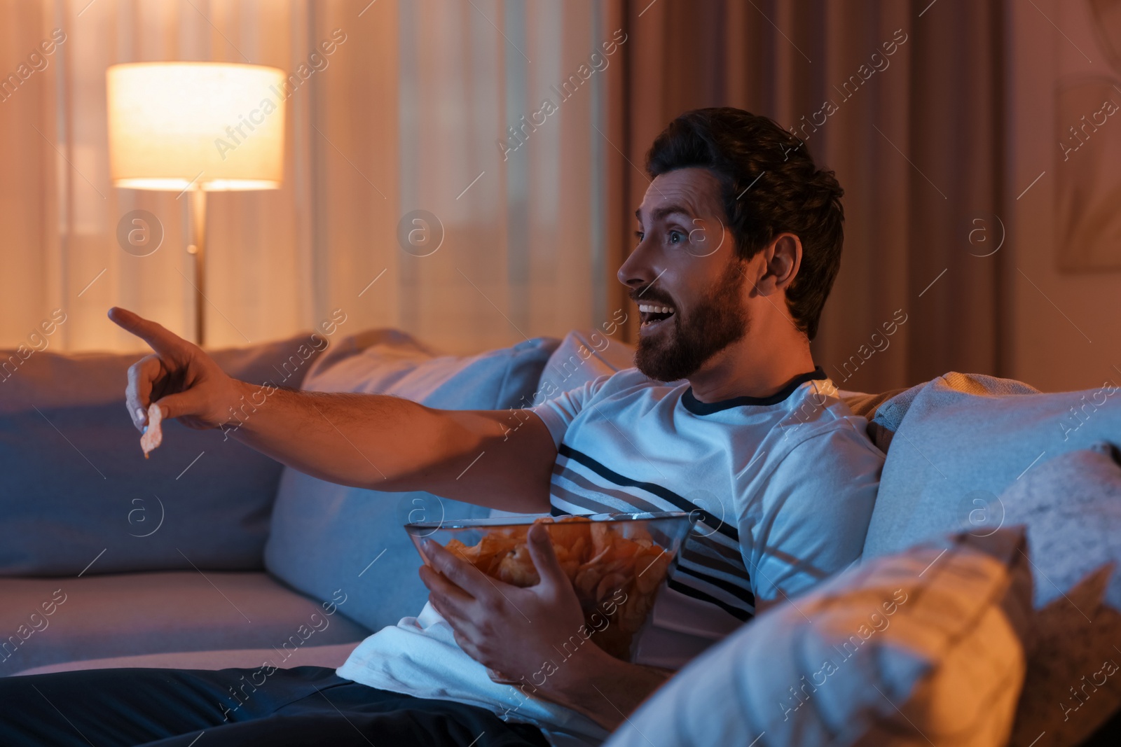 Photo of Man eating chips while watching TV on sofa at night. Bad habit