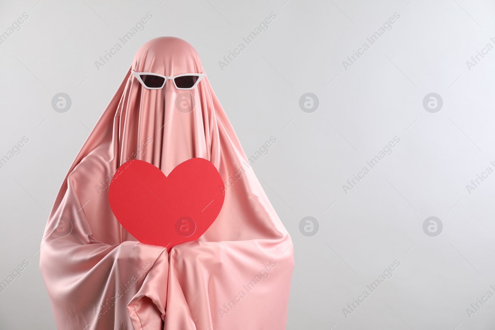 Photo of Cute ghost. Woman in pink sheet with sunglasses and red heart on light grey background, space for text