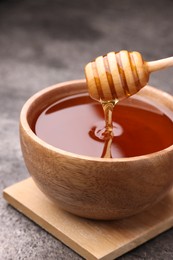 Photo of Pouring delicious honey from dipper into bowl on grey textured table, closeup