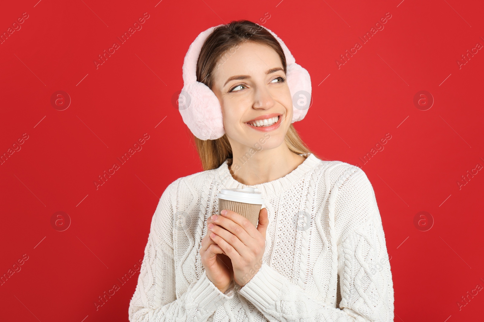 Photo of Happy woman with cup of drink wearing warm earmuffs on red background