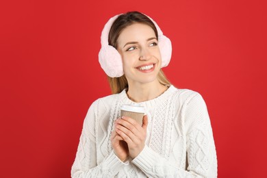 Happy woman with cup of drink wearing warm earmuffs on red background