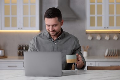 Photo of Happy man with cup of drink working on laptop at white marble table in kitchen