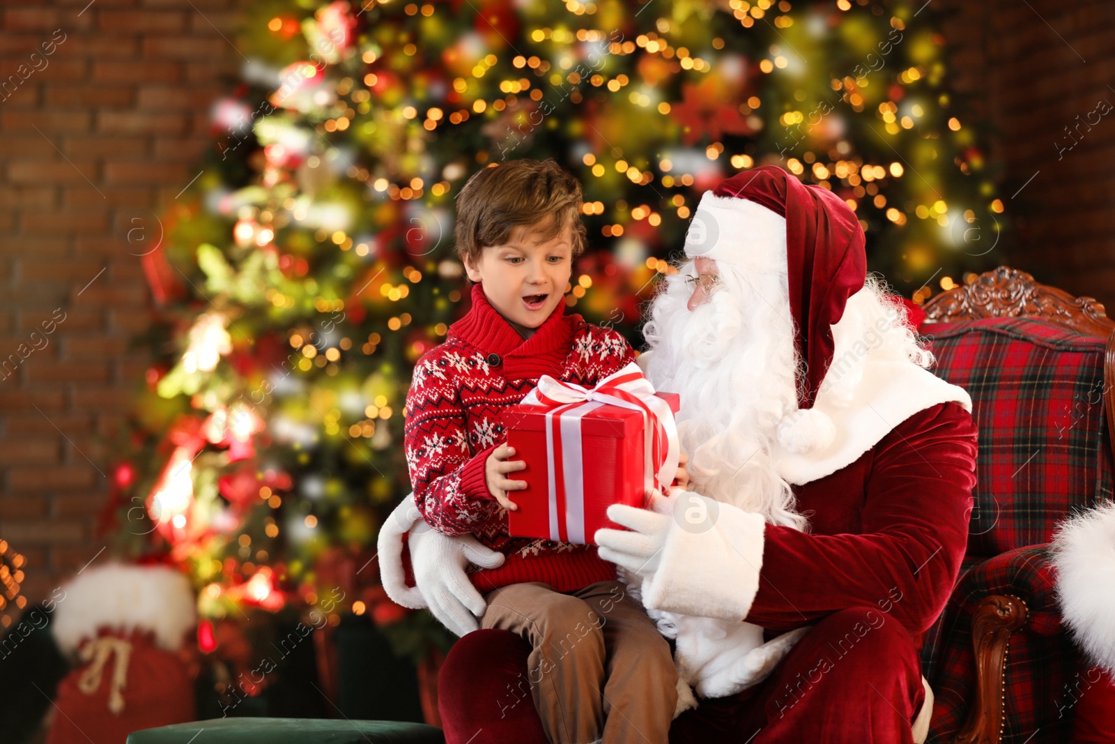 Photo of Santa Claus and little boy with gift near Christmas tree indoors