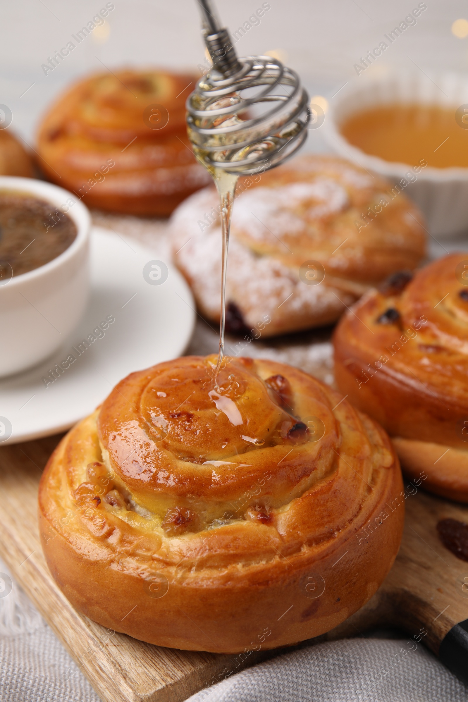 Photo of Pouring honey onto delicious rolls with raisins at table, closeup. Sweet buns