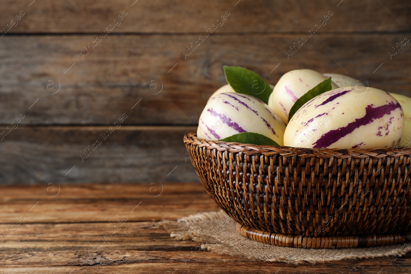 Photo of Fresh ripe pepino melons with green leaves in wicker basket on wooden table, space for text
